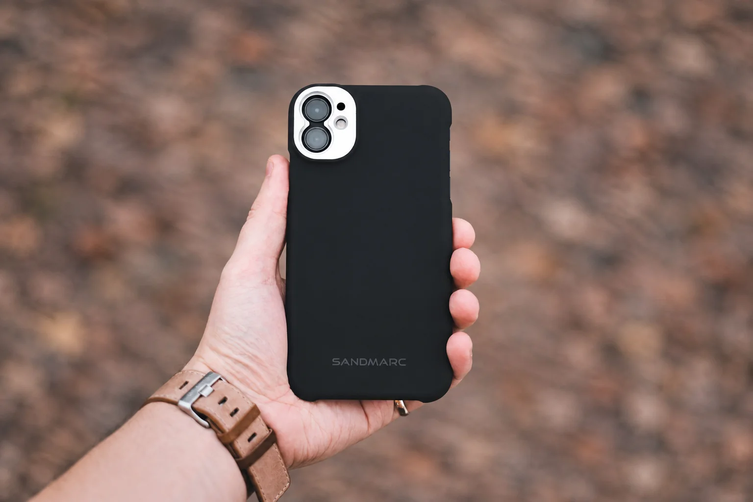 A hand with a brown watch strap holding a black protective phone case.